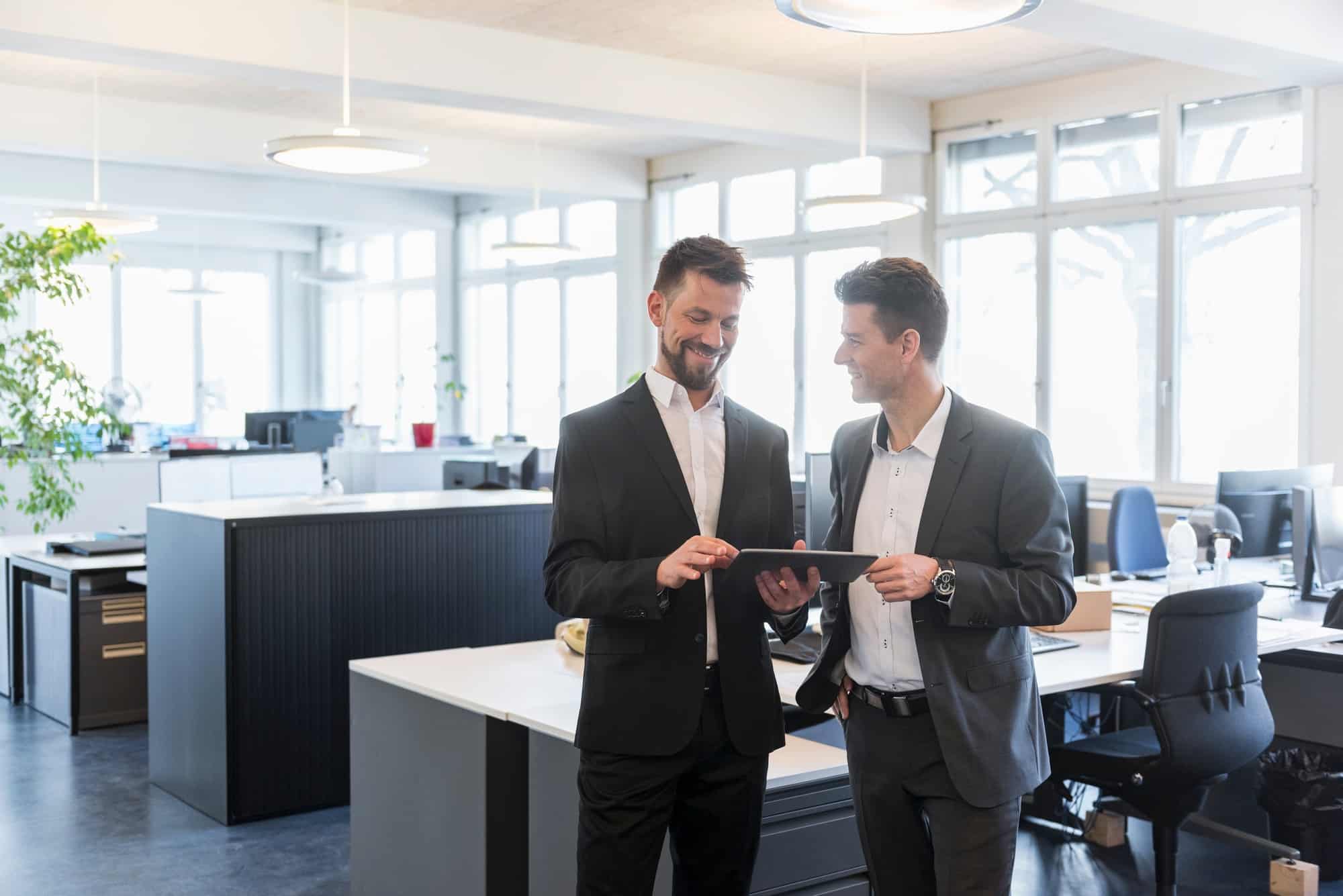Two businessmen standing in office, discussing solutions, using digital tablet